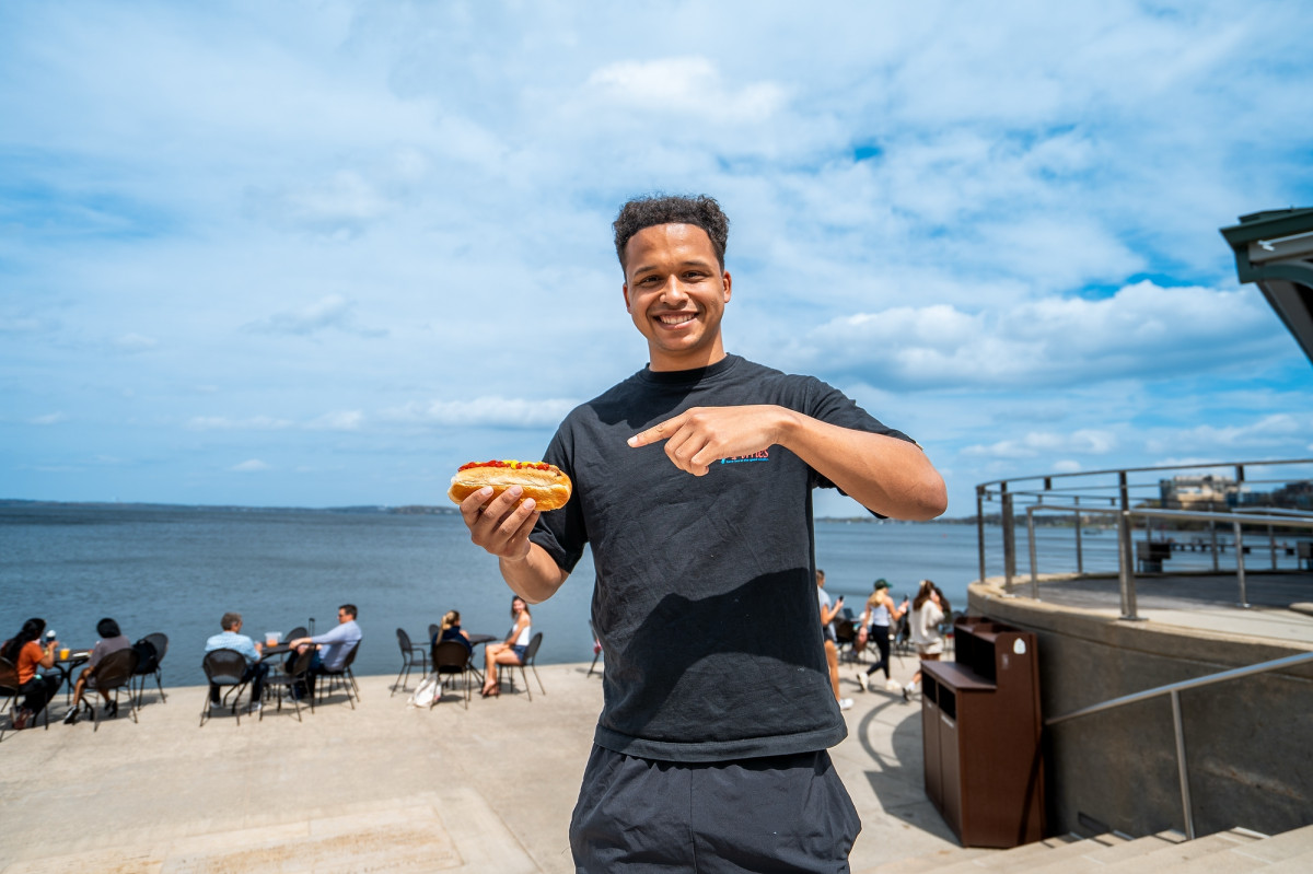 A student points at a brat in his hand in front of the lake.