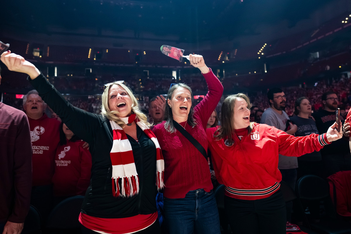 Three women with arms linked at the waist raise their free hands to sing along to the song Varsity.