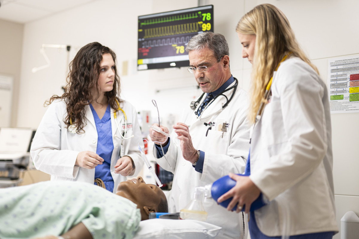 Two medical students and an instructor practice airway management on a medical simulation mannequin in a clinical training room with monitoring equipment in the background.