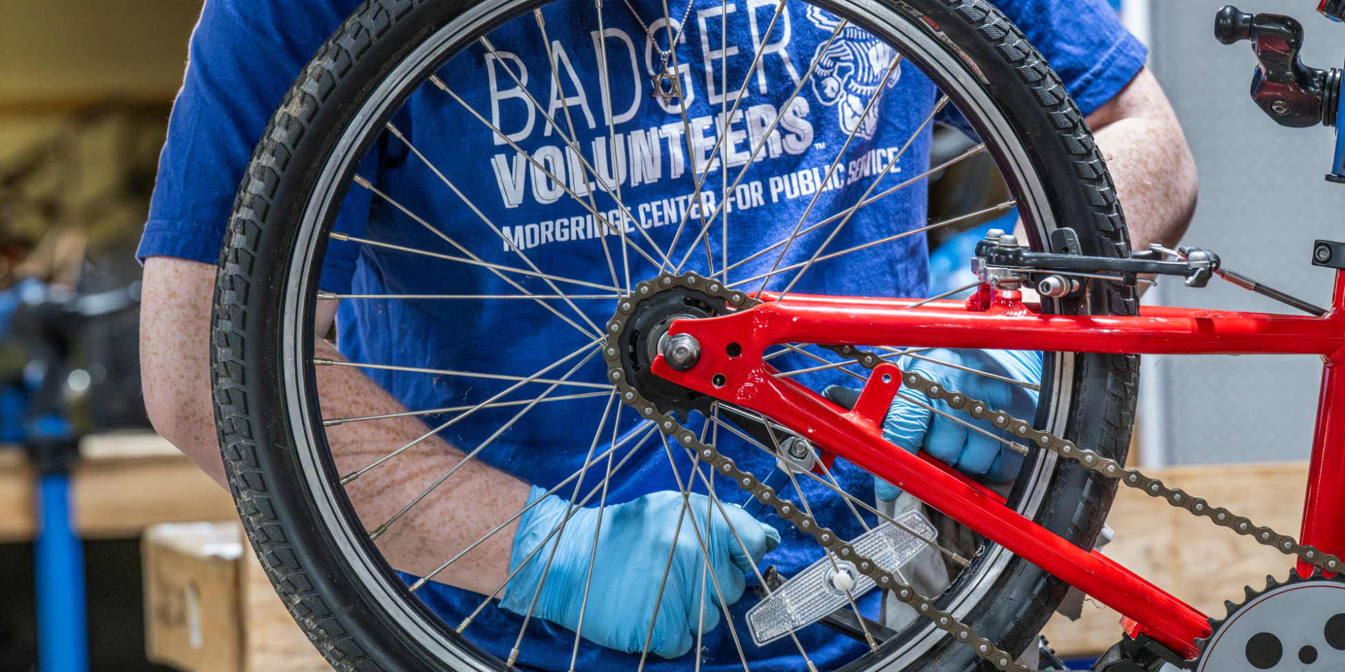 A person wearing blue gloves adjusts the rear wheel and chain of a red bicycle, with a blue shirt reading “Badger Volunteers, Morgridge Center for Public Service” visible in a workshop setting.
