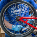 A person wearing blue gloves adjusts the rear wheel and chain of a red bicycle, with a blue shirt reading “Badger Volunteers, Morgridge Center for Public Service” visible in a workshop setting.