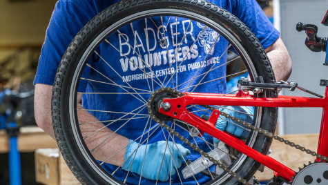 A person wearing blue gloves adjusts the rear wheel and chain of a red bicycle, with a blue shirt reading “Badger Volunteers, Morgridge Center for Public Service” visible in a workshop setting.