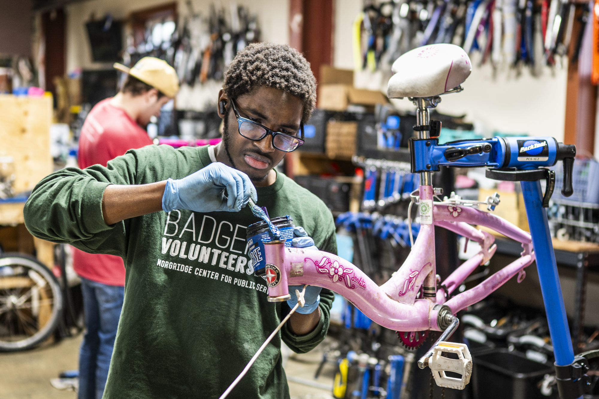 A person wearing gloves and a green 'Badger Volunteers' shirt works on a small pink bicycle mounted on a repair stand in a busy workshop.