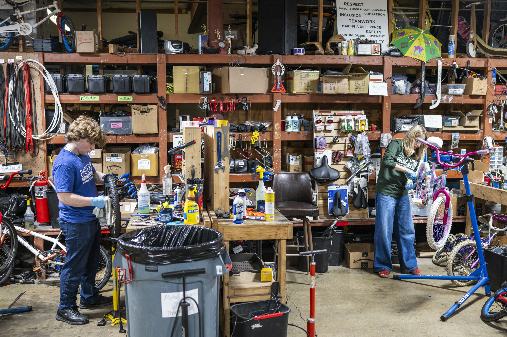 Two people repair bicycles at workstations inside a large workshop filled with shelves of tools, supplies, and bike components, with bikes in various stages of repair.