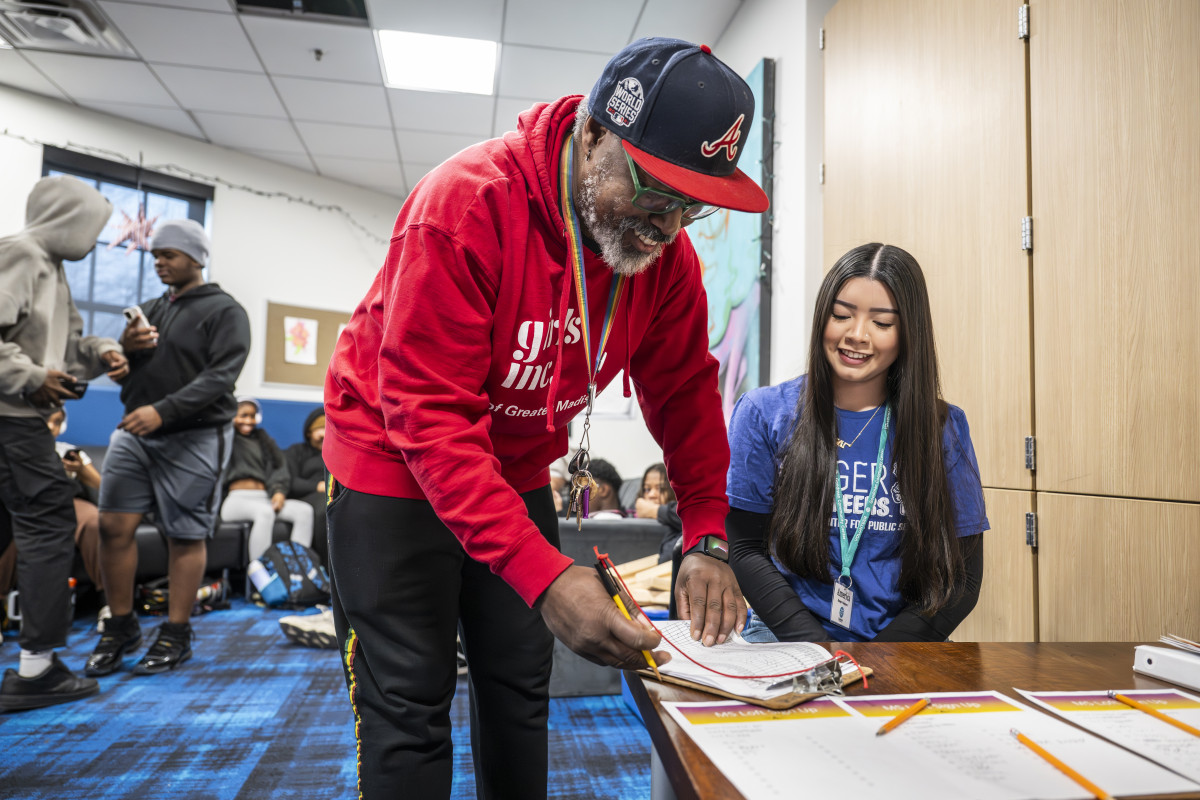 A person leans over a table to write on a sign-up board next to a Badger volunteer, with middle schoolers nearby in a community room.