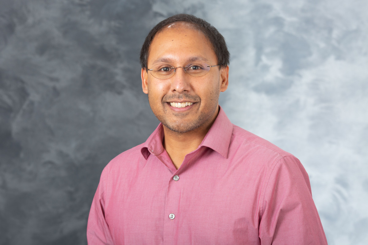 Studio portrait photo of a man smiling to the camera. He's wearing rimless glasses and a reddish pink button down shirt.