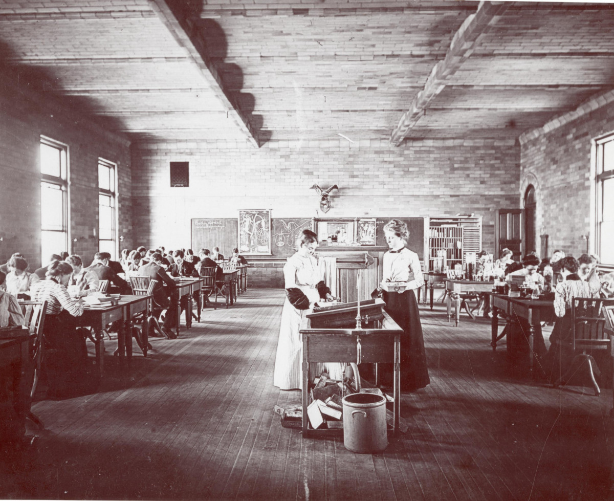 A black and white photograph showing students working in a laboratory. The room is brightly lit. Two female students stand in the center of the room around what appears to be a sink while male and female students work seated at lab benches.