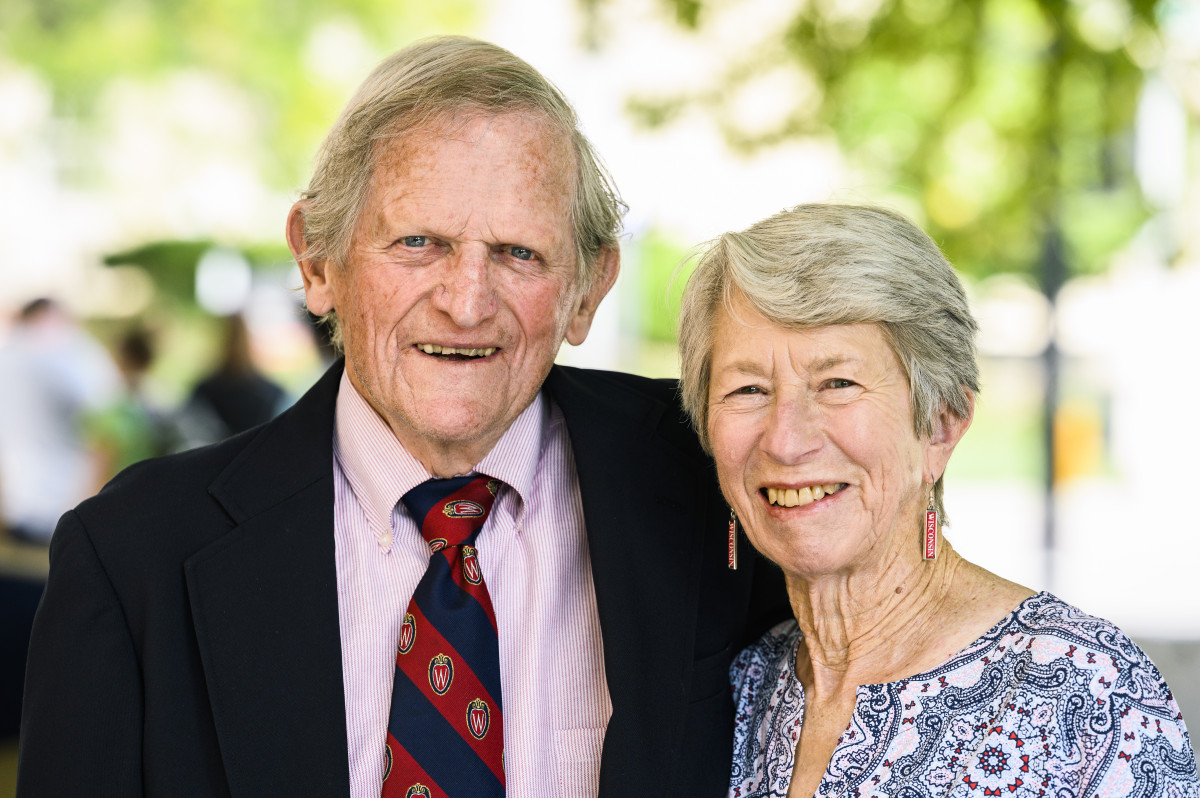 A gray-haired man in a striped tie and a gray-haired woman in a blue patterned dress stand together smiling.