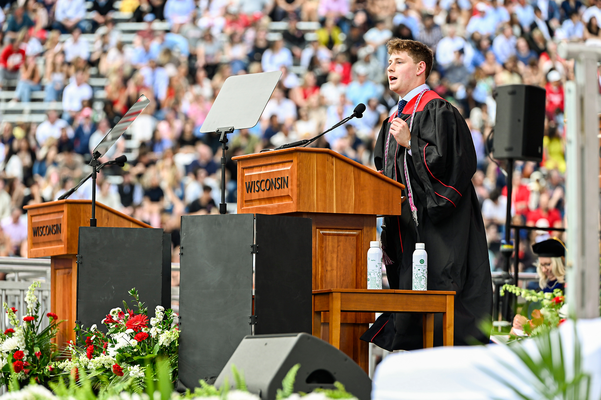 Schwartz wears a gradation cap and gown and stands behind a podium as he addresses the crowd. 