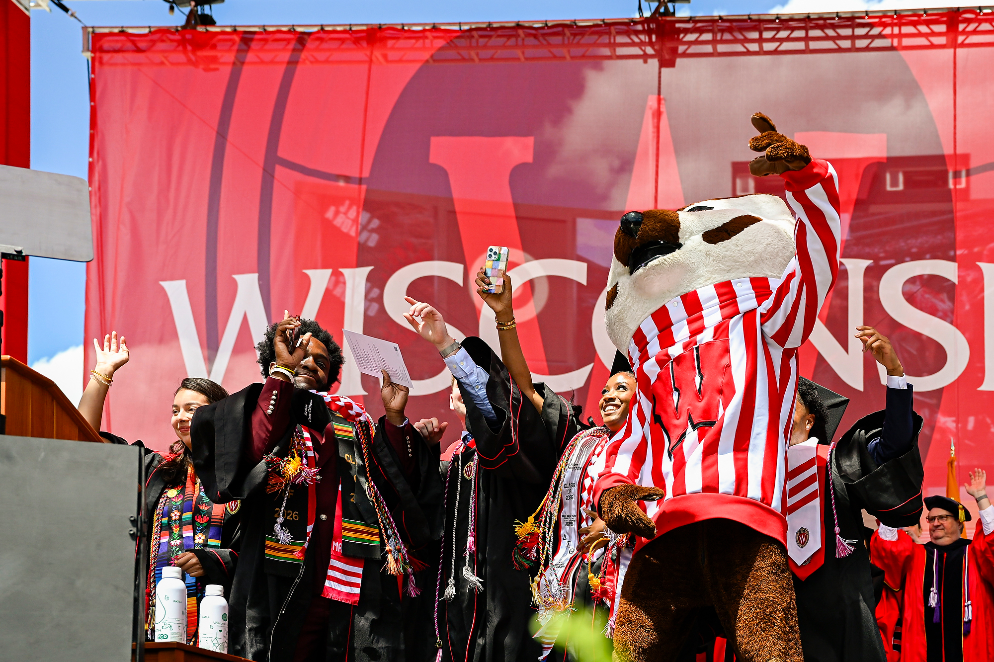 A group of students all wearing commencement robes stand on stage with Bucky Badger and dance in celebration.