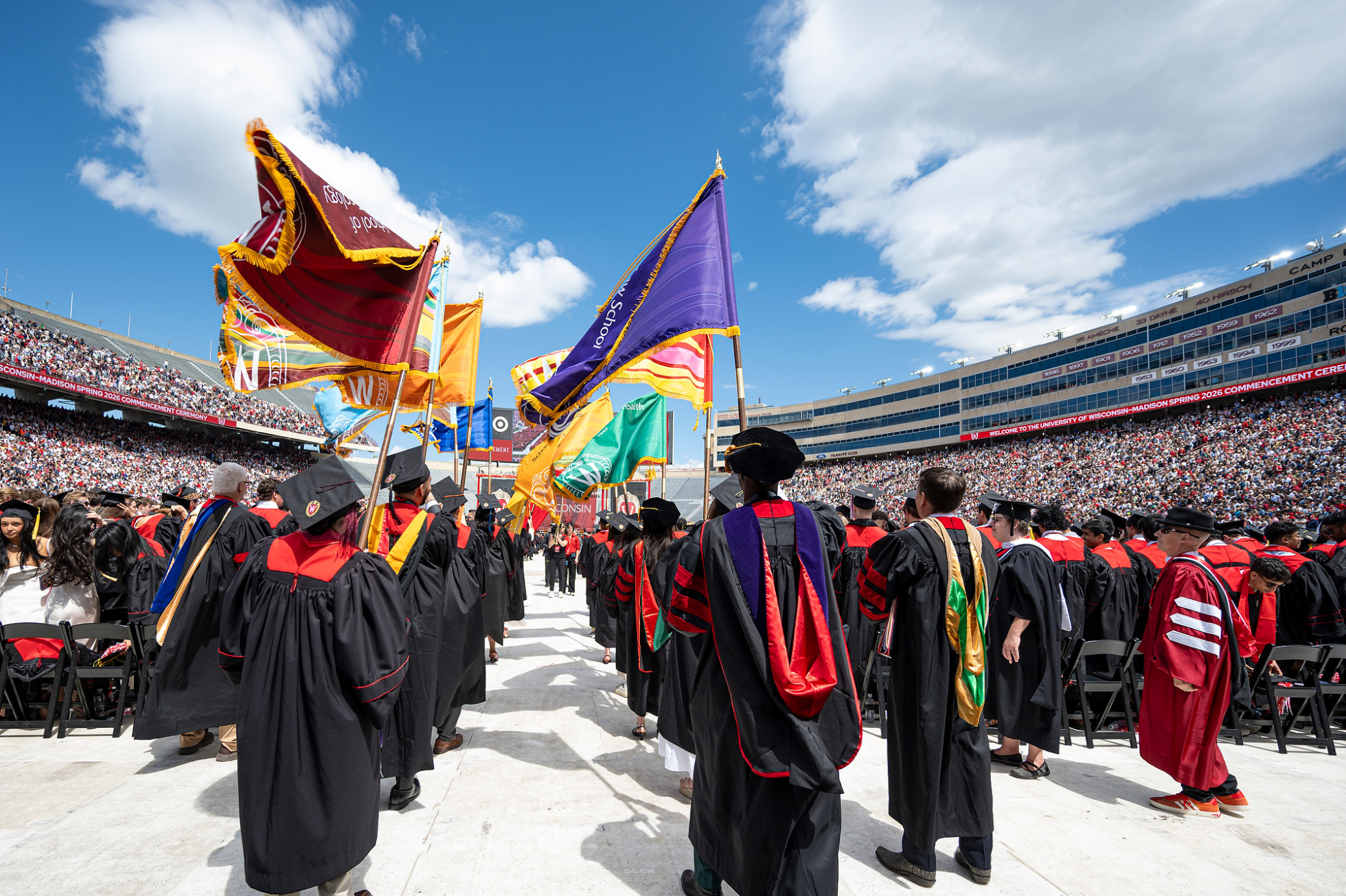 Flag bearers in caps and gowns walk down the center aisle of Camp Randall stadium, surrounded by graduates and cheering spectators.