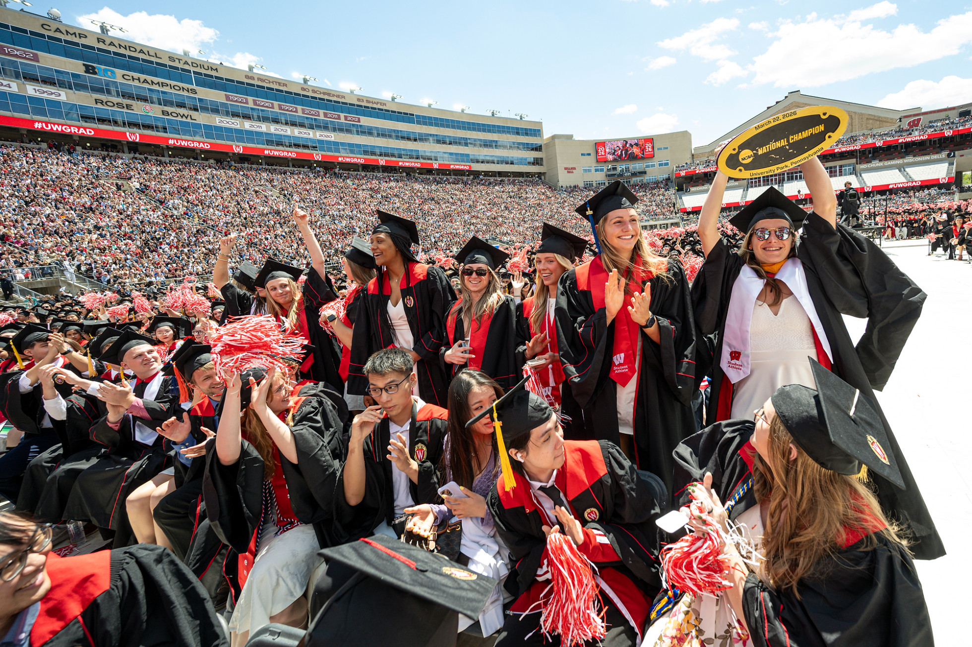 Wearing their graduation regalia, members of the Women’s UW Hockey Team stand and cheer, with one holding up a National Championship trophy.