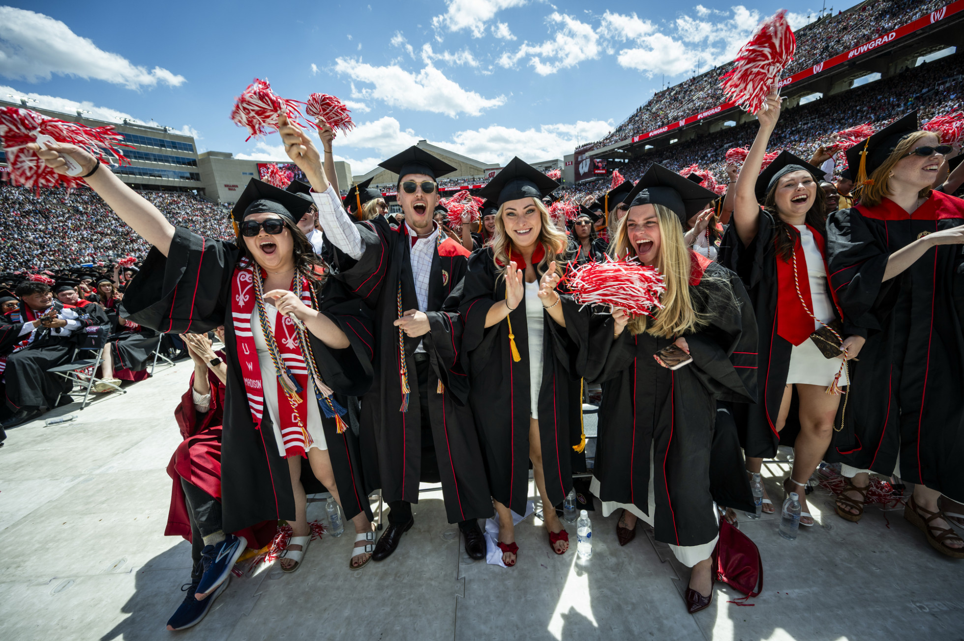 A group of enthusiastic graduates wearing their caps and gowns raise red and white pom-poms and jump in celebration.
