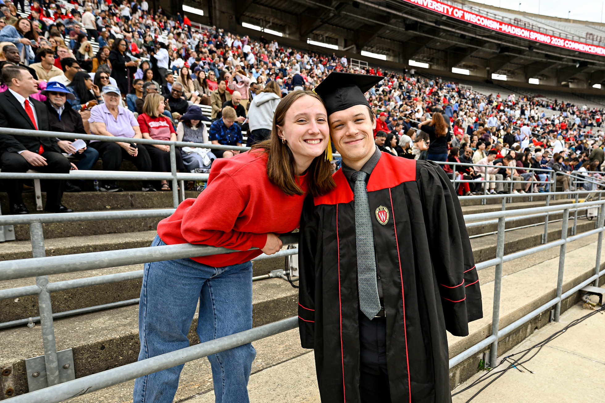 A smiling Porter wears a graduation cap and gown and stands next to Beeman who leans over the seating railing to join him for the picture. 