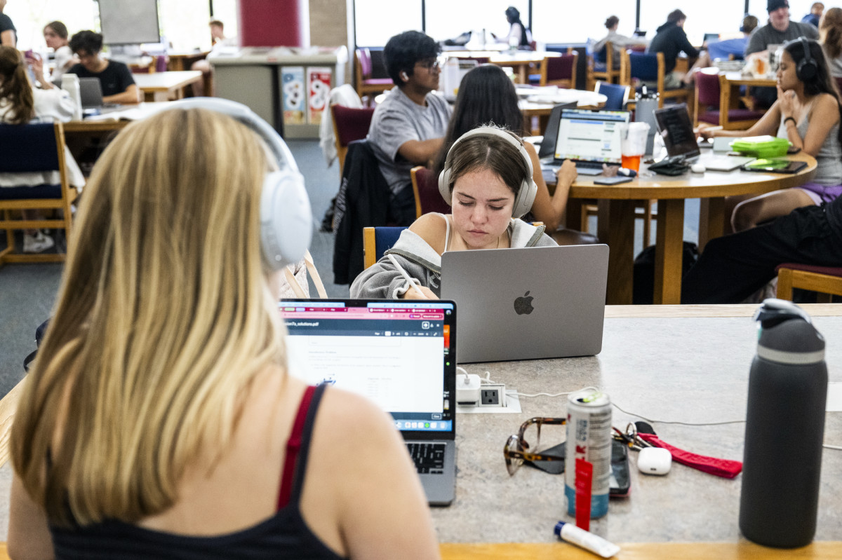 Busy study area with people seated at shared tables using laptops and notebooks. In the foreground, a person wearing over‑ear headphones writes on a laptop with a stylus. Water bottles, chargers, and personal items rest on the table.