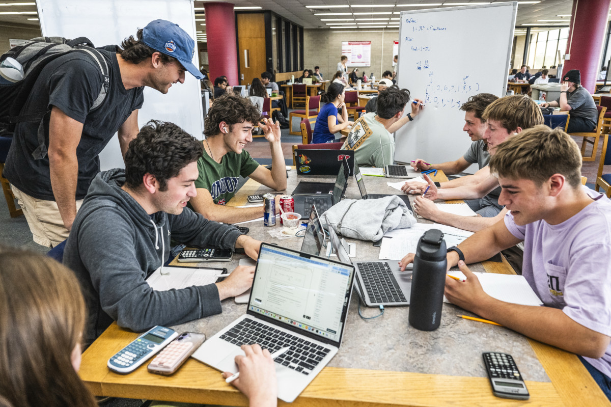 A group of students study together with laptops open at a table in a crowded library common area. One student is standing and smiling as looks over another's shoulder at his laptop.