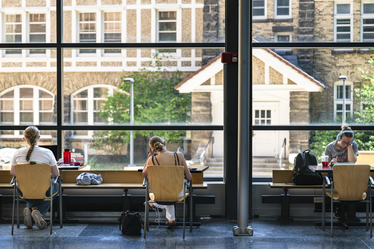 A serene study scene with students sitting far apart at cafe tables in front of a floor-to-ceiling glass window that looks out onto a Tudor-style limestone building.