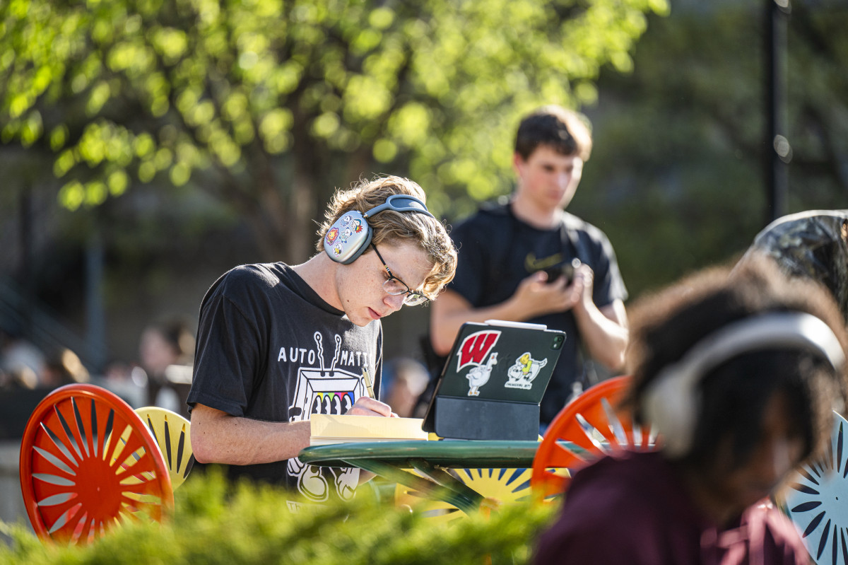 A student with headphones on takes paper notes while sitting outdoors at a metal table on a sunny terrace. His headphones and laptop cover are full of stickers, including a red UW W.