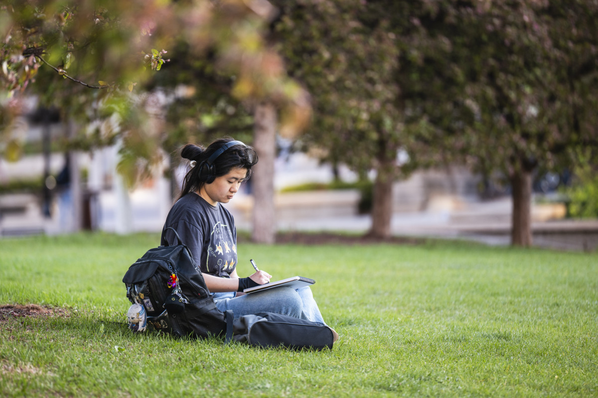 A student sits by herself on a grassy lawn listening to headphones and taking notes on paper.
