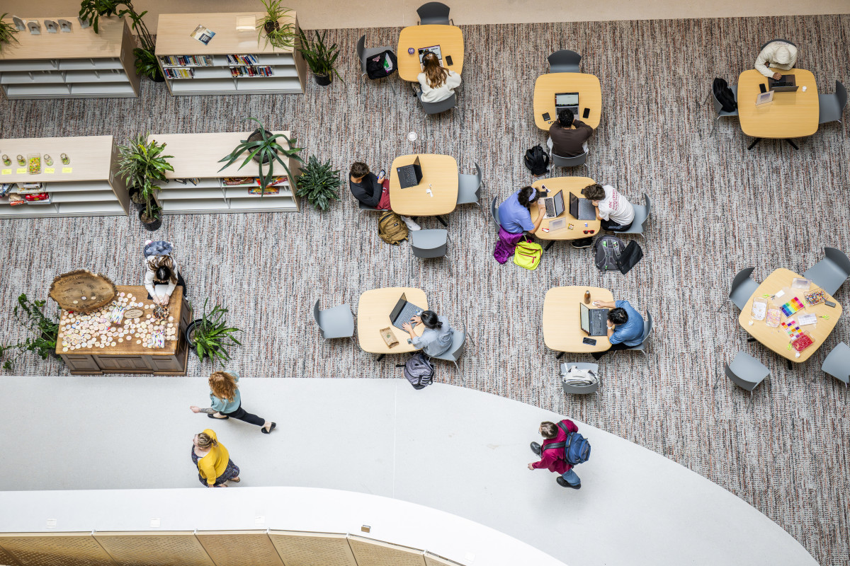 An overhead view of students sitting at small desks in an open area. Some students are walking in a walkway.