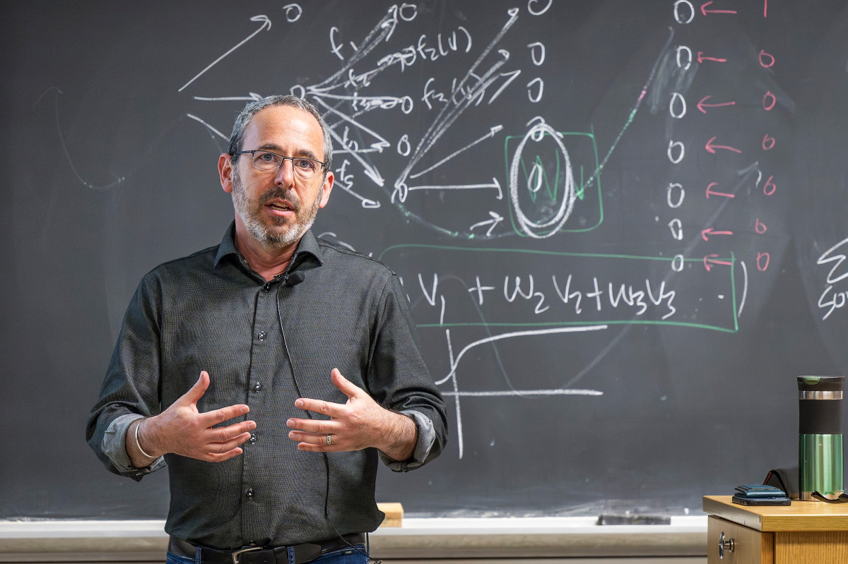 A man in a dark gray button down shirt stands in front of a blackboard covered in equations. He is speaking and gesturing with his hands.