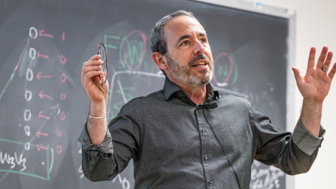 A man in a dark gray button down shirt stands in front of a blackboard covered in equations. He is speaking and gesturing with his hands.
