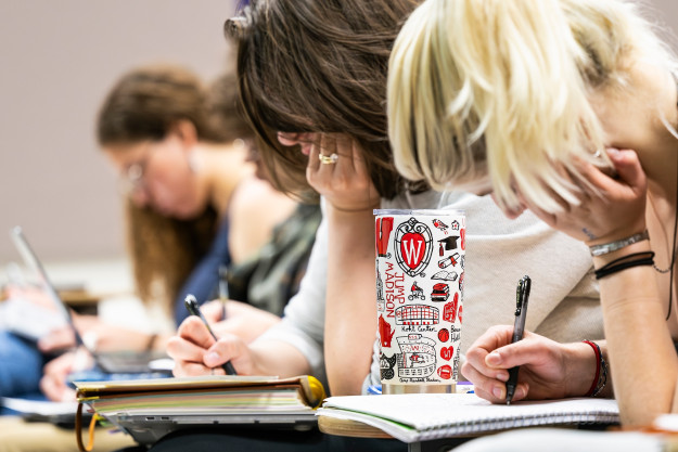 Two students taking notes in class have the same posture: heads down, face resting on their palms as they write paper notes.