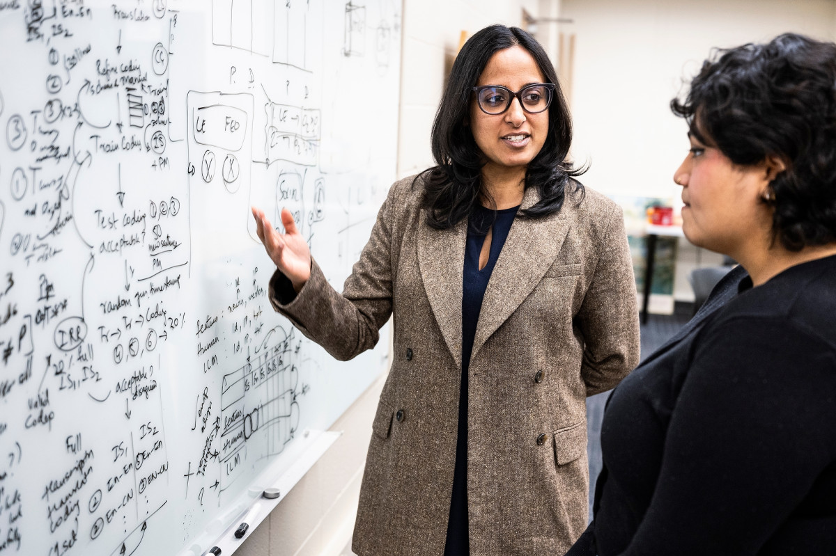 Shamya Karumbaiah stands in front of a whiteboard talking to an undergraduate student in the classroom. 