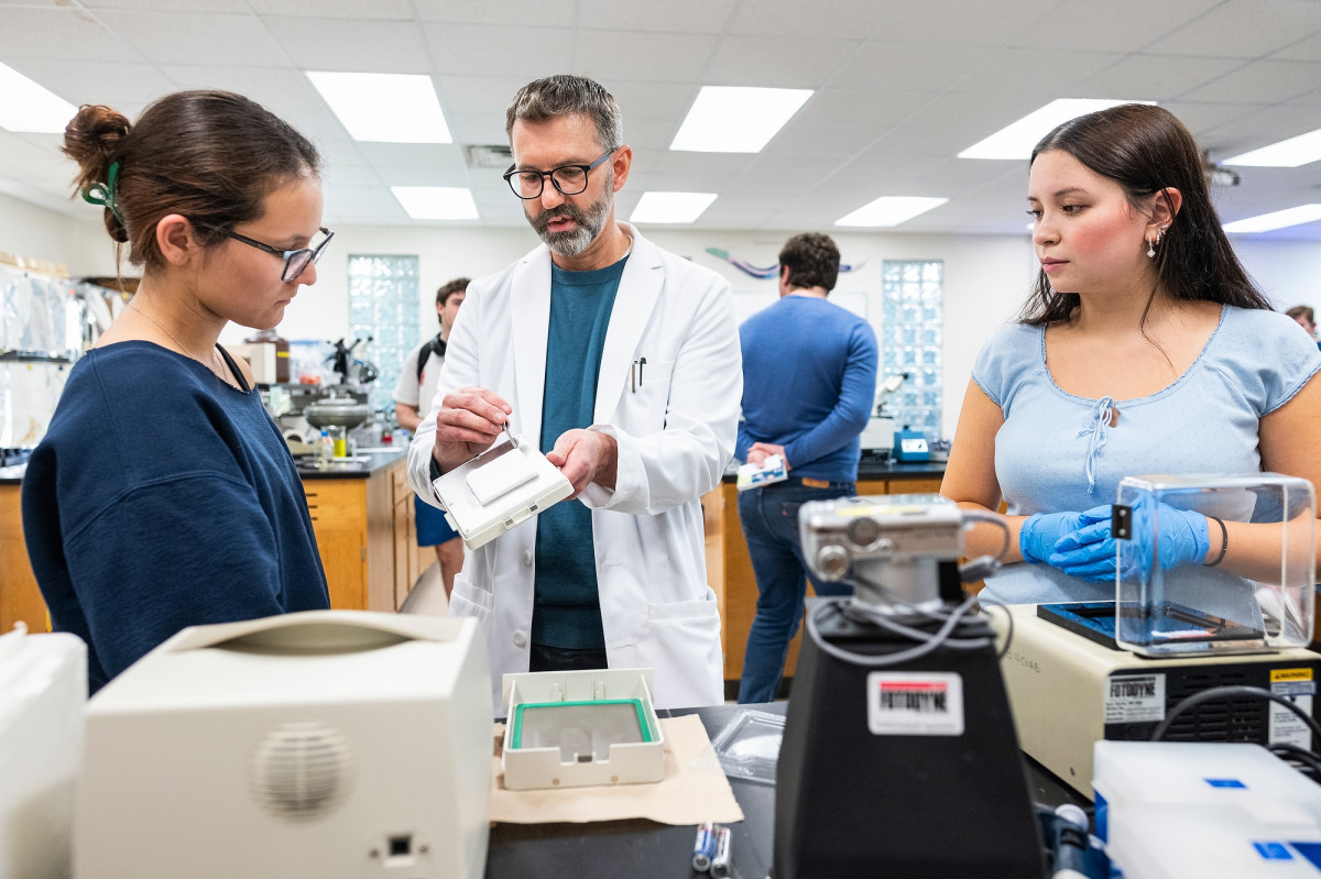 Seth McGee, wearing a white lab coat, shows two undergraduate students a piece of testing equipment.