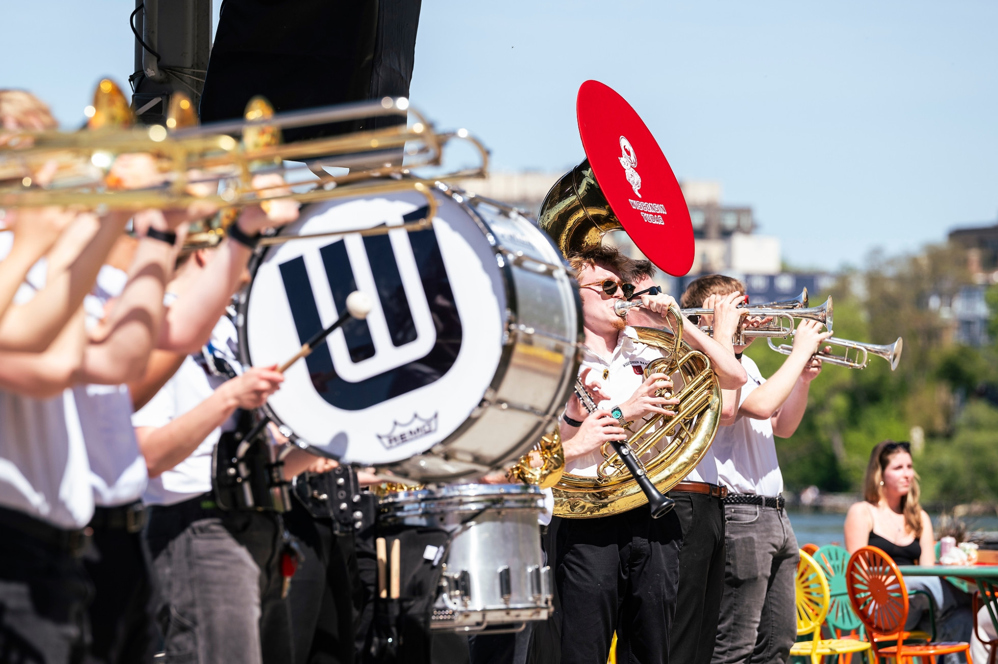 A close up photos shows the line of UW marching band members playing their instruments against the backdrop of Terrace chairs and Lake Mendota.