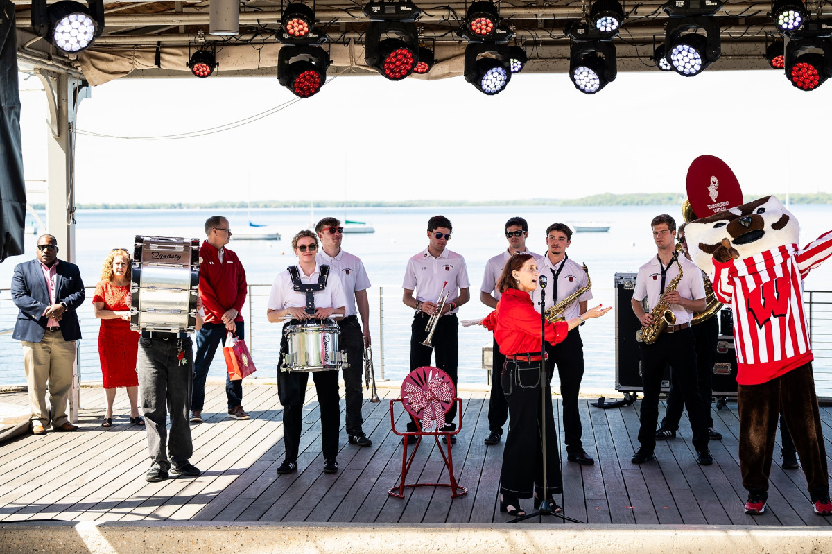 Bogost stands on a stage next to Bucky Badger as she speaks to the crowd through a standing microphone. Behind her the UW marching bands stands in a line with their instruments.