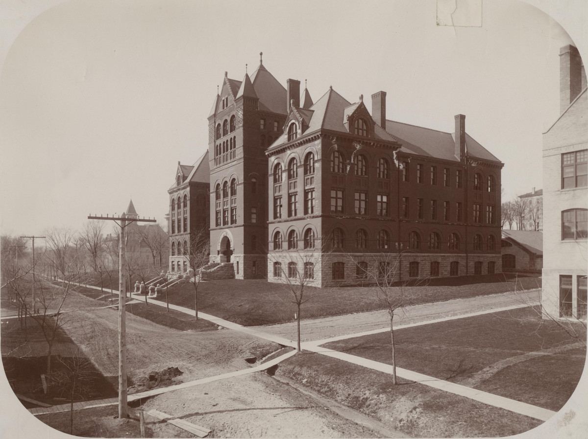 A black and white photograph of a large brick building at the corner of intersecting dirt roads.