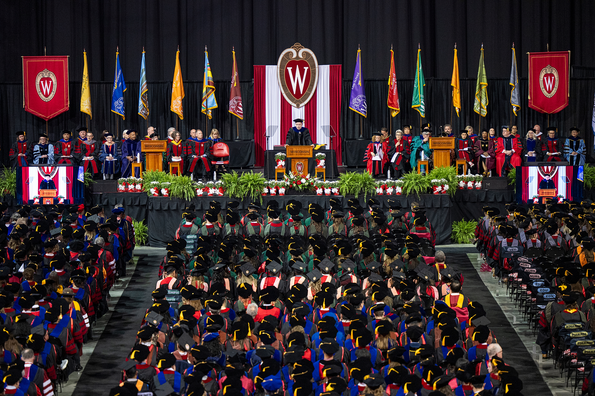 Shown from the crowd’s perspective, Provost John Zumbrunnen speaks at the podium on the commencement stage, flanked by seated members of the official party. 