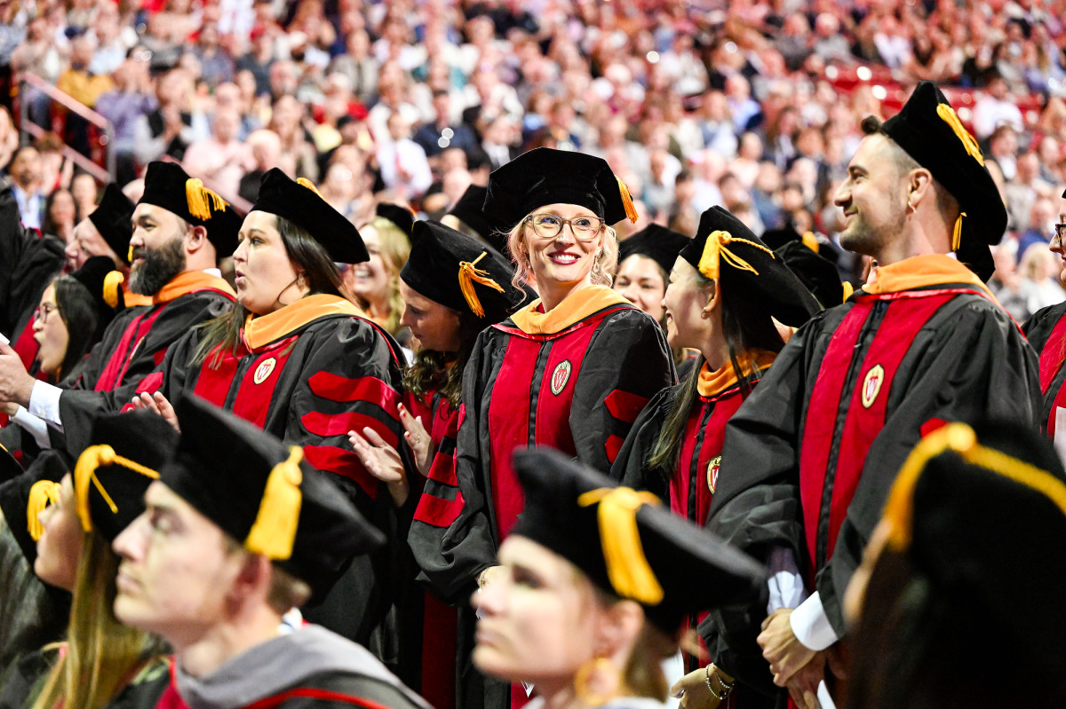A crowd of smiling graduates, wearing commencement regalia, stand and applaud.