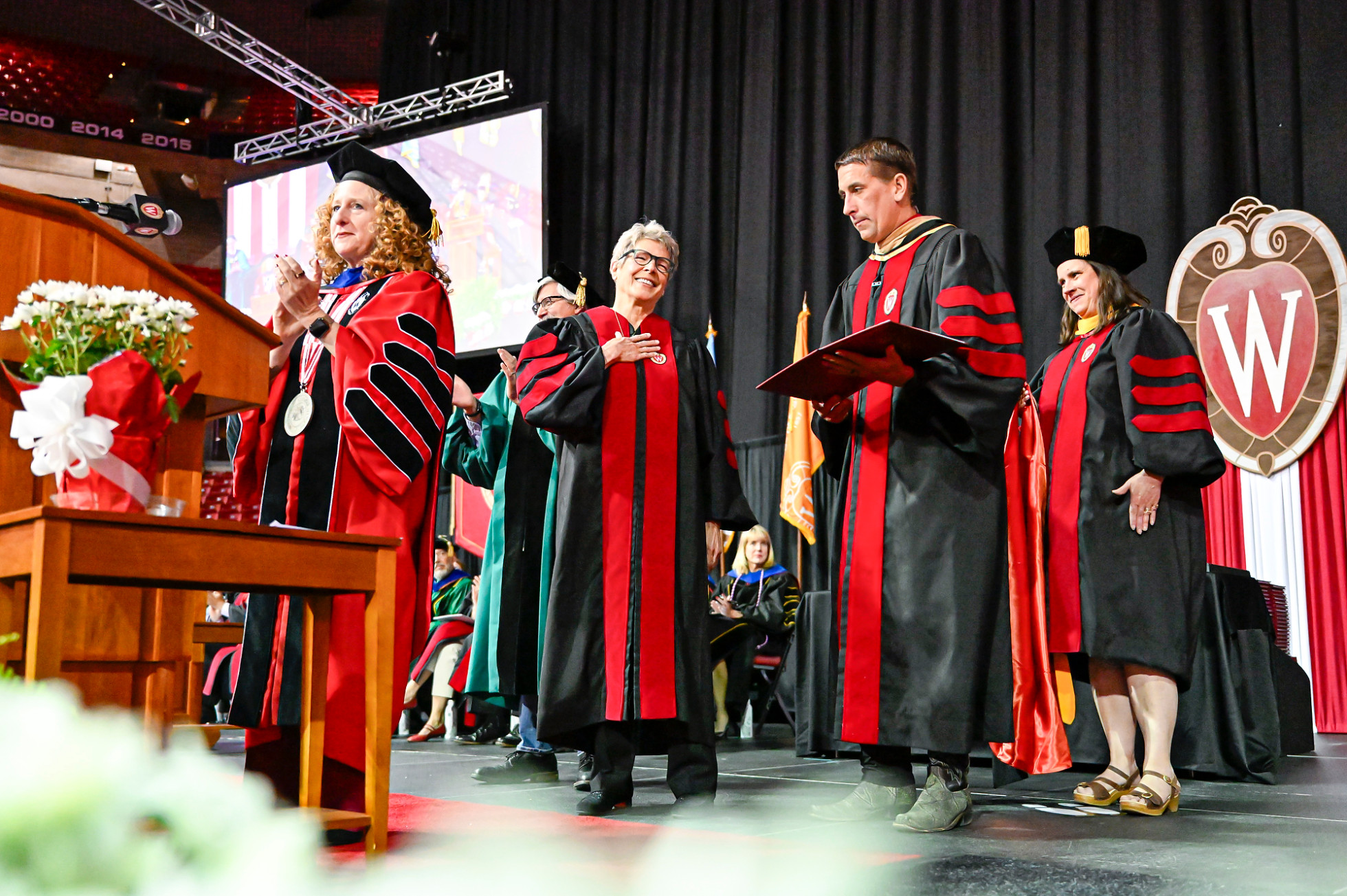 Tia Nelson, wearing a commencement gown, smiles and places a hand over her heart on stage, flanked by Chancellor Jennifer Mnookin and other members of the official party.