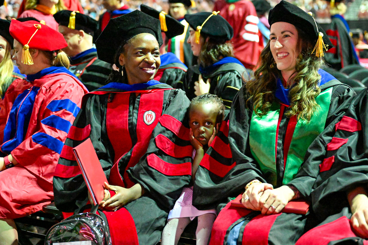 Graduate Dorothy Lsoto, wearing commencement regalia and smiling while holding a diploma cover, sits next to her daughter Avia. 