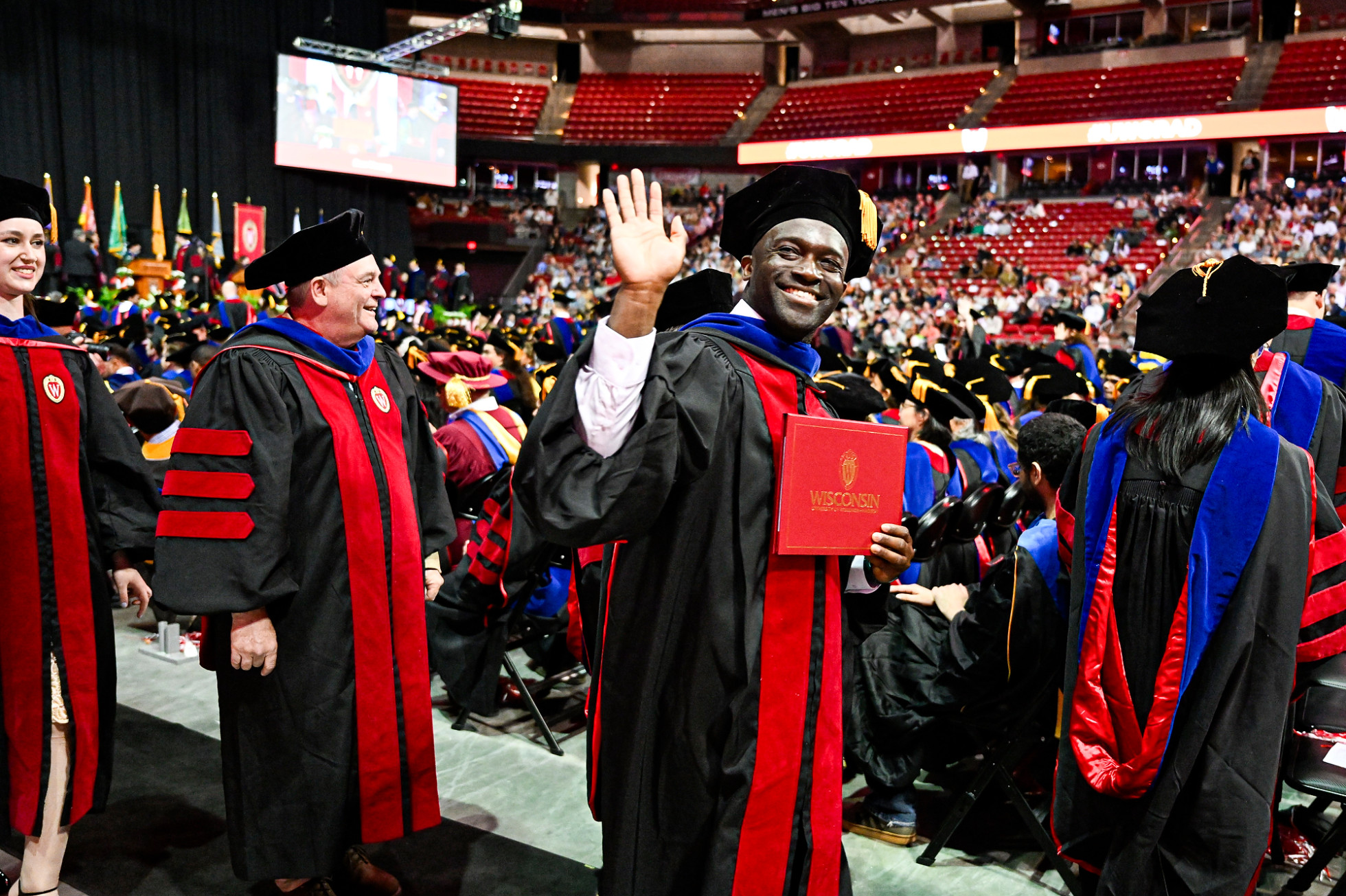 Graduate Matthew McKetty, wearing commencement regalia, smiles and waves at the crowd while walking down the aisle and holding his diploma cover. 
