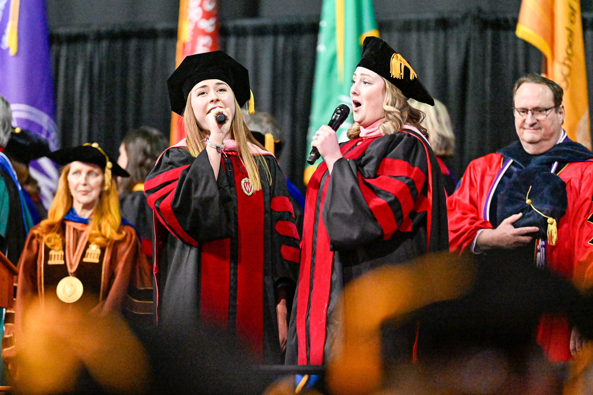 Madison Barrett and Eloise Berkley, wearing commencement regalia, sing into handheld microphones with members of the official party in the background.