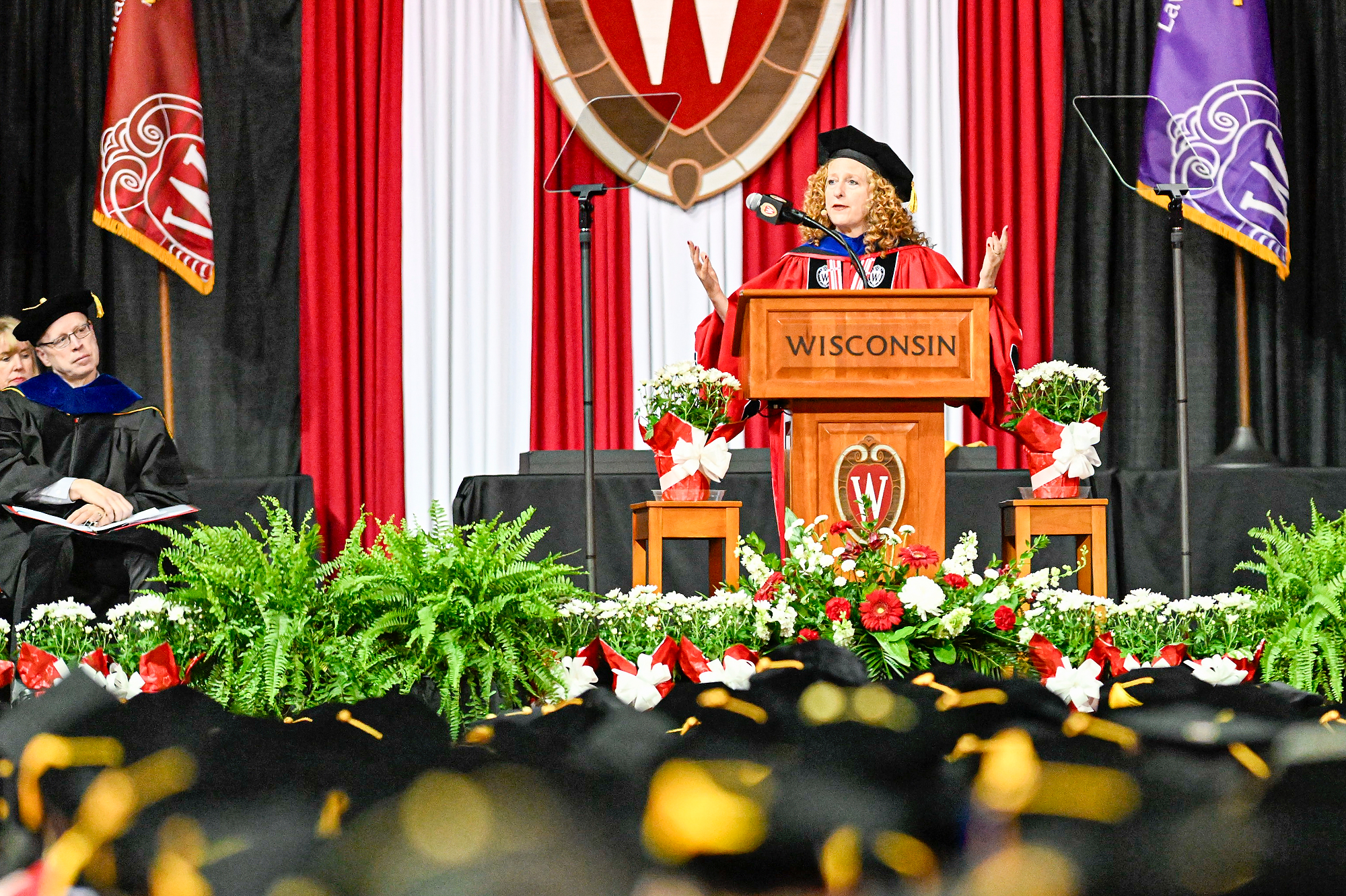 Chancellor Jennifer Mnookin, wearing commencement regalia, speaks at the podium with her hands raised on the commencement stage. 