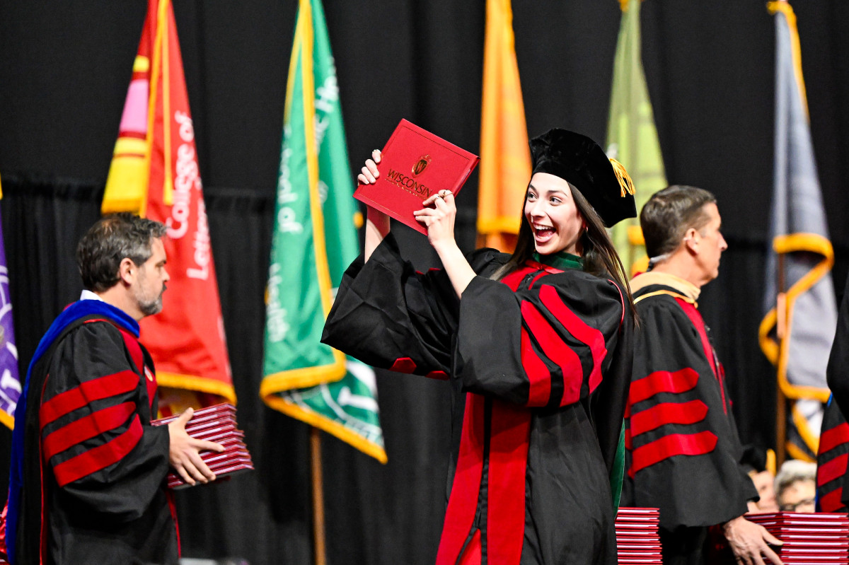 A graduate, wearing commencement regalia, smiles and poses with her degree cover while walking across the stage.