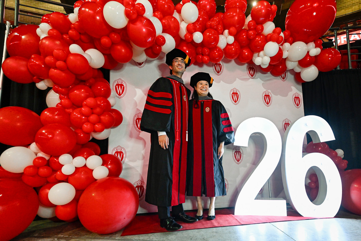 Graduates Ethan Elazegui and Deborah Liu pose for a photo in front of red and white balloon arch and a UW-branded backdrop. 