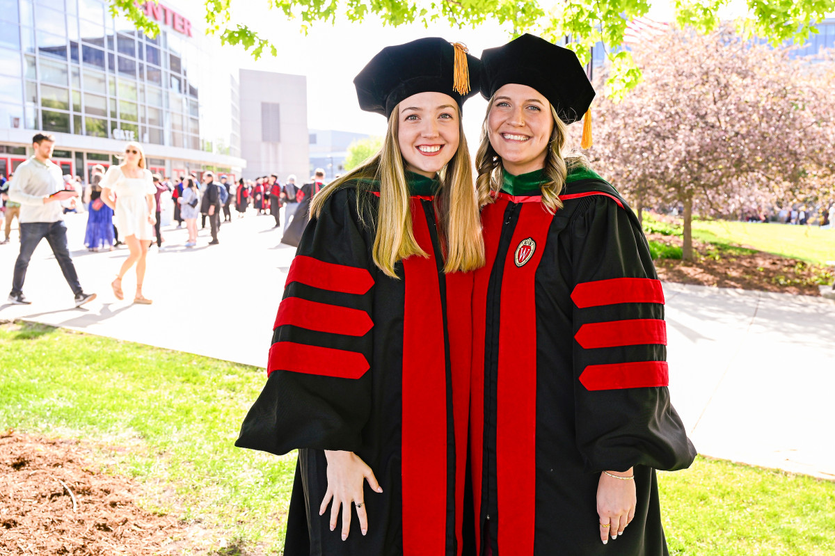 Graduates Lauren Hlubek and Sierra Block, wearing commencement regalia, pose for a photo outside of the Kohl Center. 