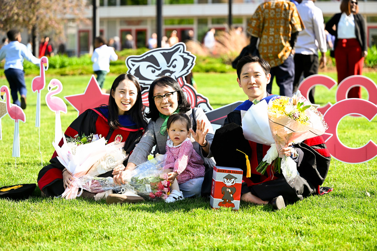 A family, including two graduates wearing commencement regalia and holding flowers, sit on the Kohl Center lawn and smile for the camera. 