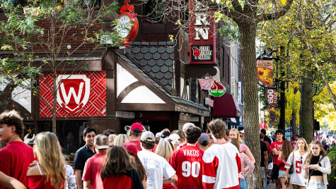 A crowd wearing red and white fills the sidewalk outside State Street Brats with festive signs and fall foliage overhead.