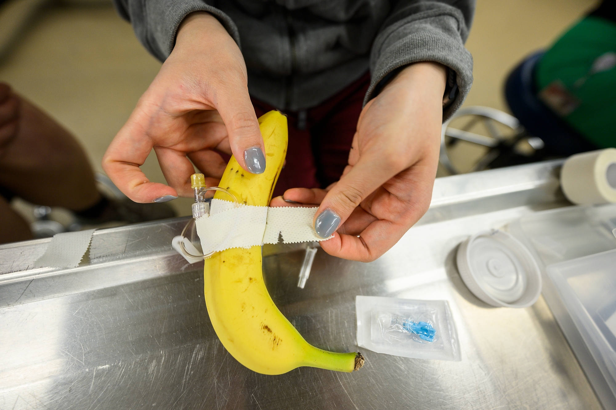 A close up photo shows a student's hands wrapping medical tape around a banana to hold a plastic catheter in place.