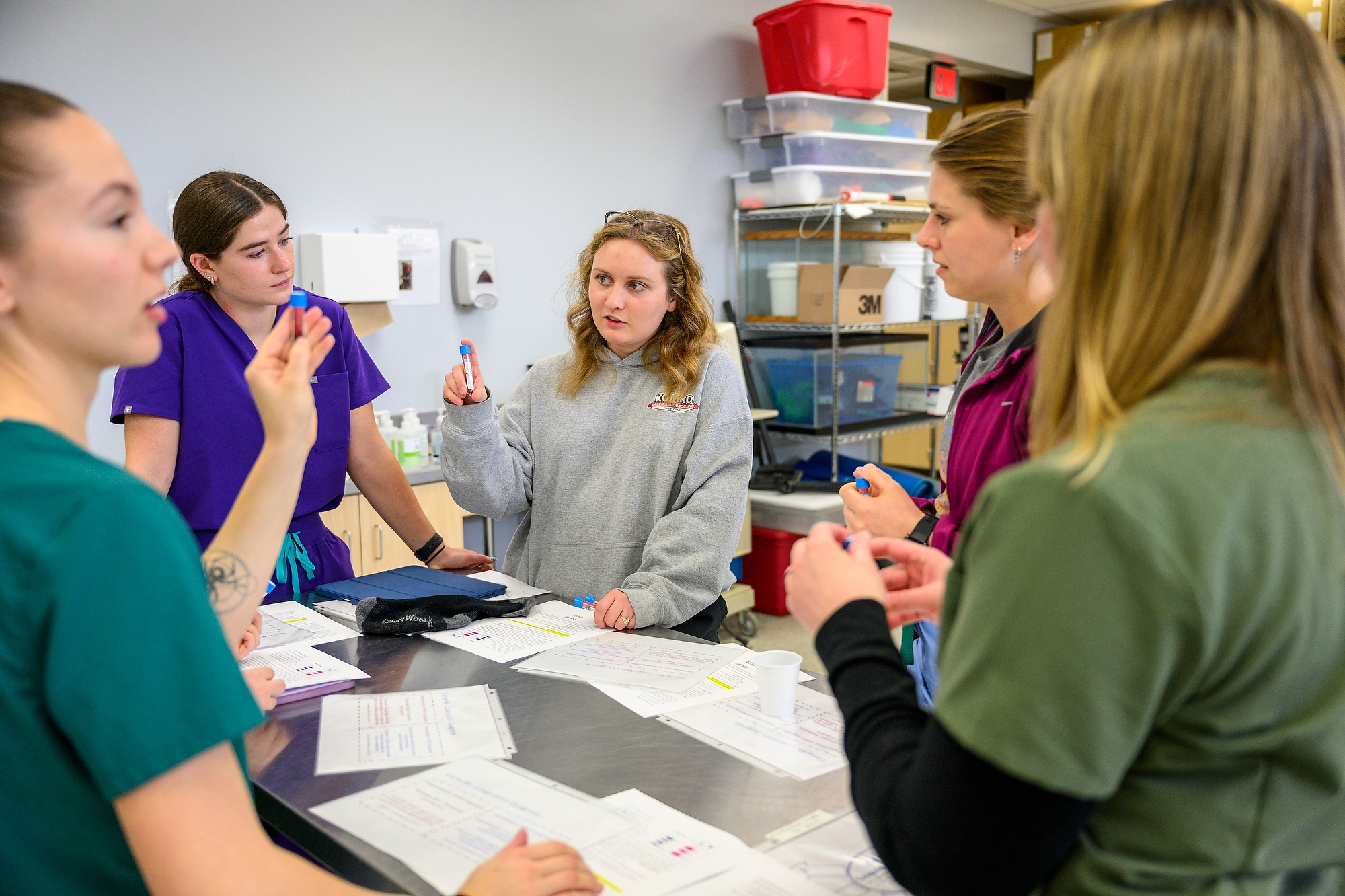 Students stand around a table holding vials of blood while discussing their lab exercise.