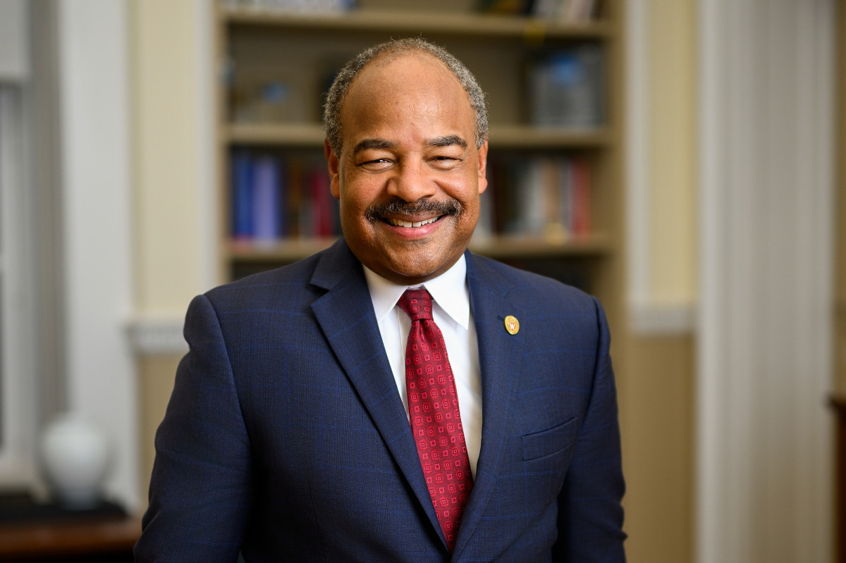 A man in a blue suit and red tie stands smiling in front of a bookcase.