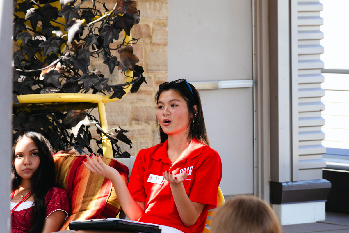 A student wearing a bright Badger red shirt sits in a yellow Terrace chair and talks to fellow students. 