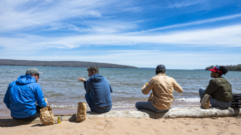 Four people sit on a log on a beach eating from brown bag lunches.