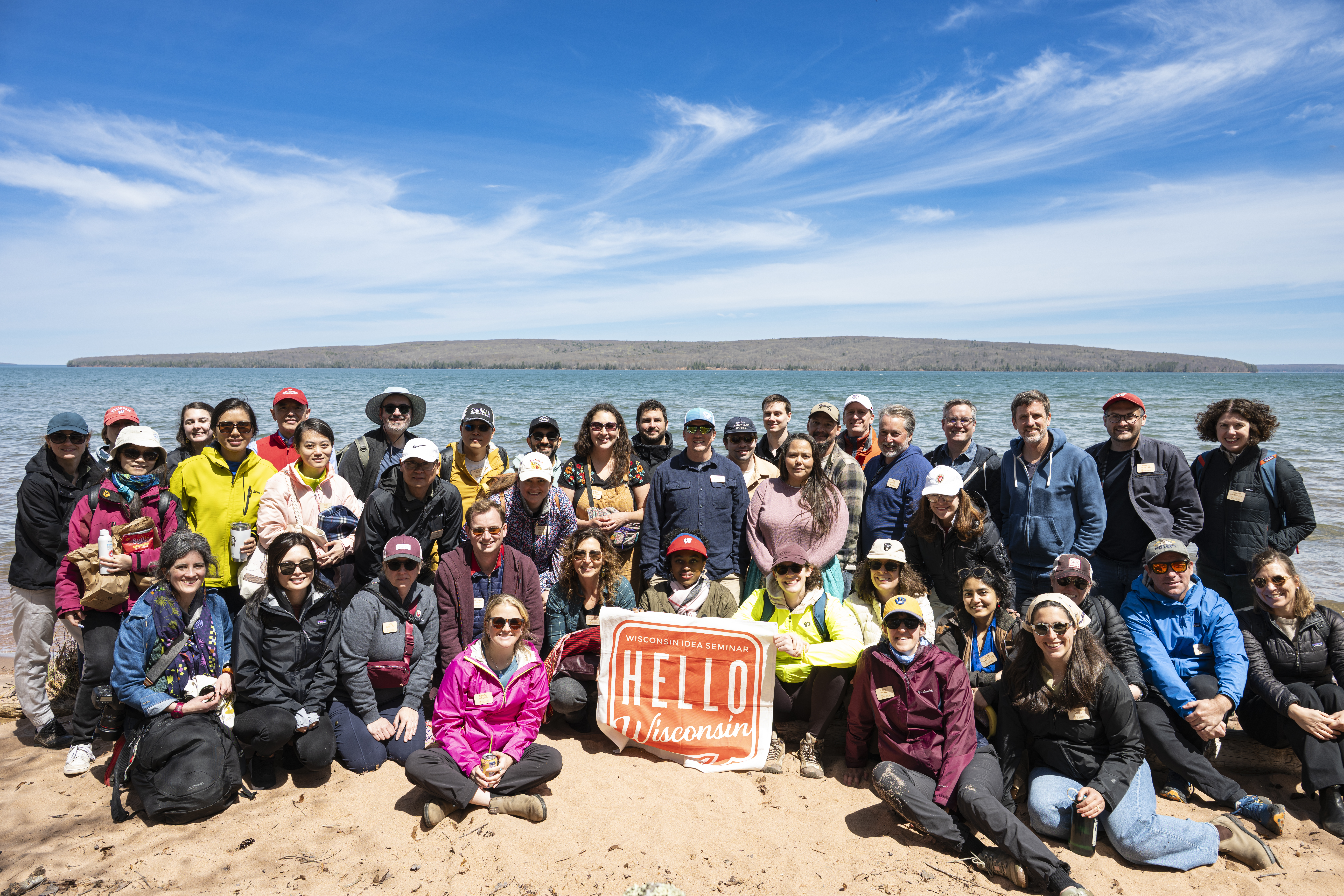 The members of the 2026 Wisconsin Ideas Seminar tour pose together in front of a large body of water with a sign that reads Hello Wisconsin.